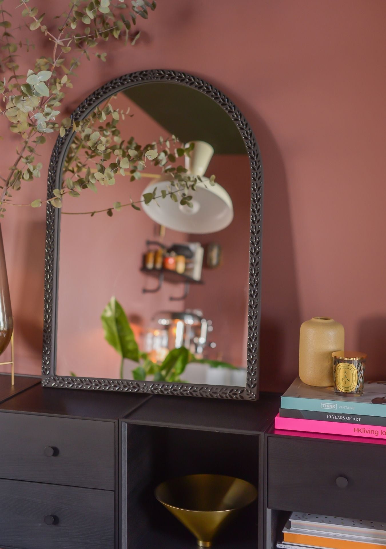 Handmade arched mosaic mirror by Pure Reflection, styled on a black cabinet against a warm terracotta wall with natural branches, candles, and books.