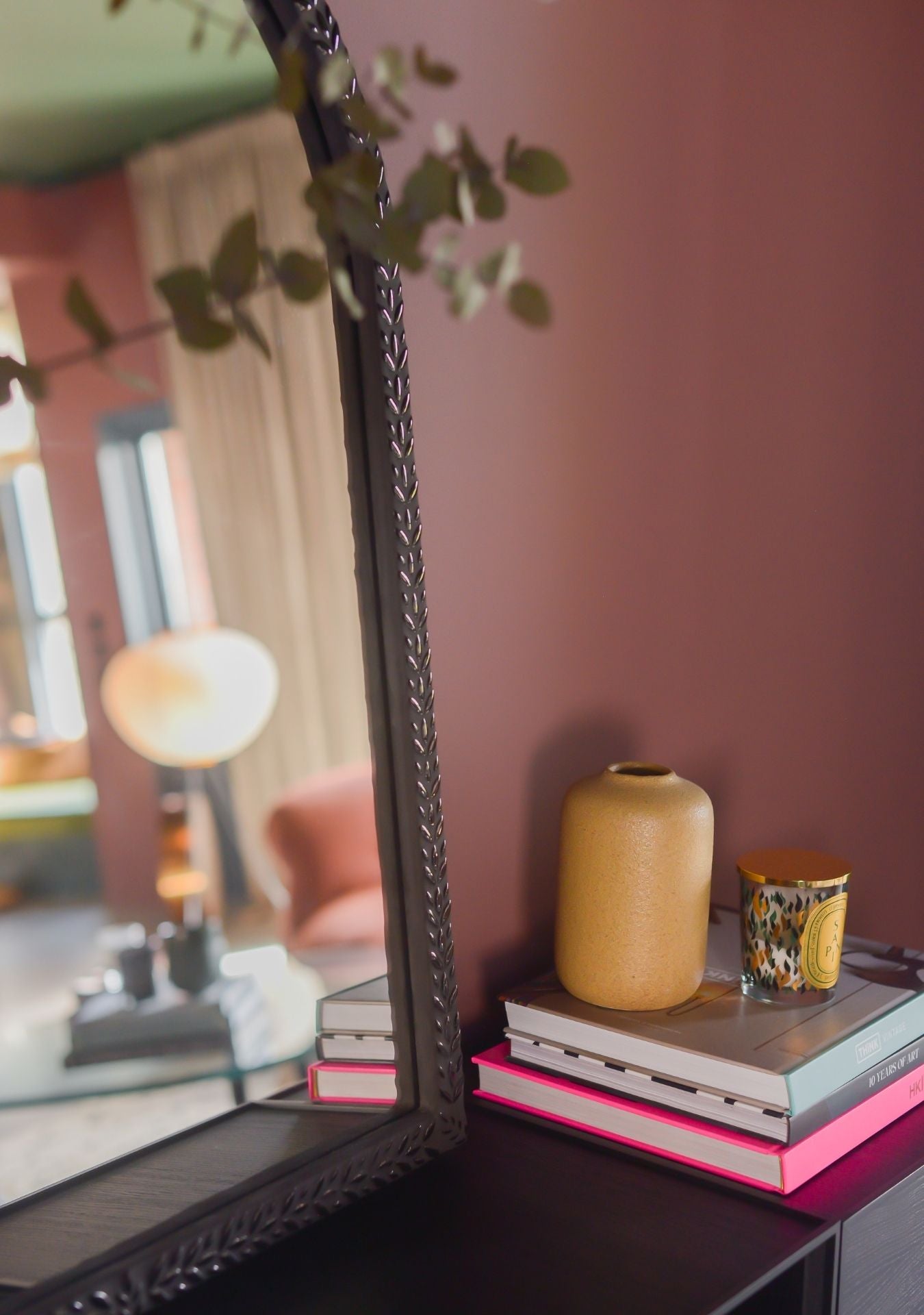 Close-up of a handmade mosaic mirror by Pure Reflection, showing detailed craftsmanship and soft reflections. Styled with a gold vase, candle, and stacked books against a warm wall.
