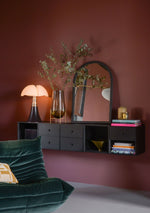 A handmade mosaic mirror from Pure Reflection displayed in a warm, elegant interior. The mirror hangs above a black floating cabinet against a terracotta wall, styled with decorative vases, books, and a sculptural lamp. A green velvet chair adds a touch of luxury to the cozy setting.