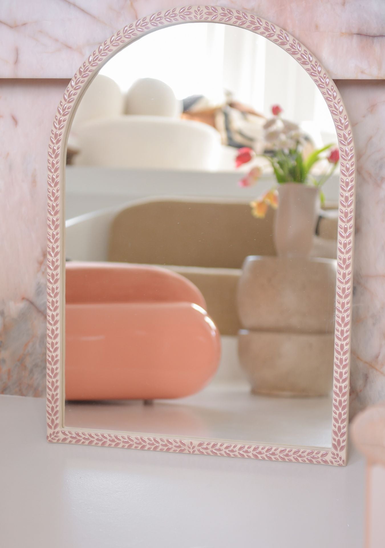 Handmade pink arched mosaic mirror by Pure Reflection, featuring a delicate leaf-patterned frame and styled in a soft, modern interior with warm neutral tones and sculptural decor pieces.