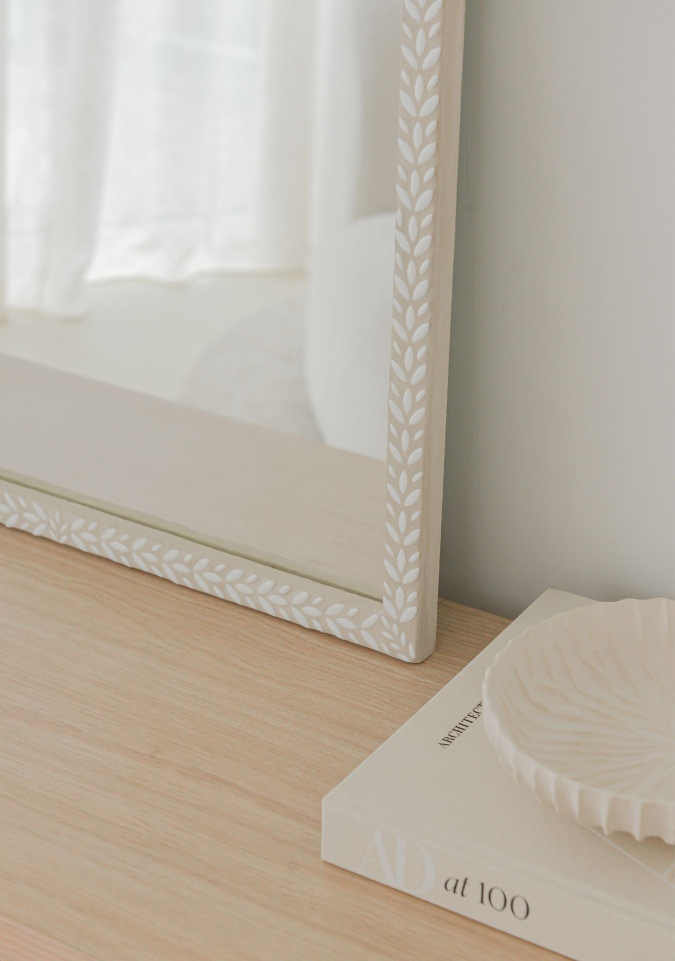 Close-up of a handmade white mosaic mirror by Pure Reflection, showing the delicate leaf-patterned frame detail on a light wooden surface in a bright, minimal interior.