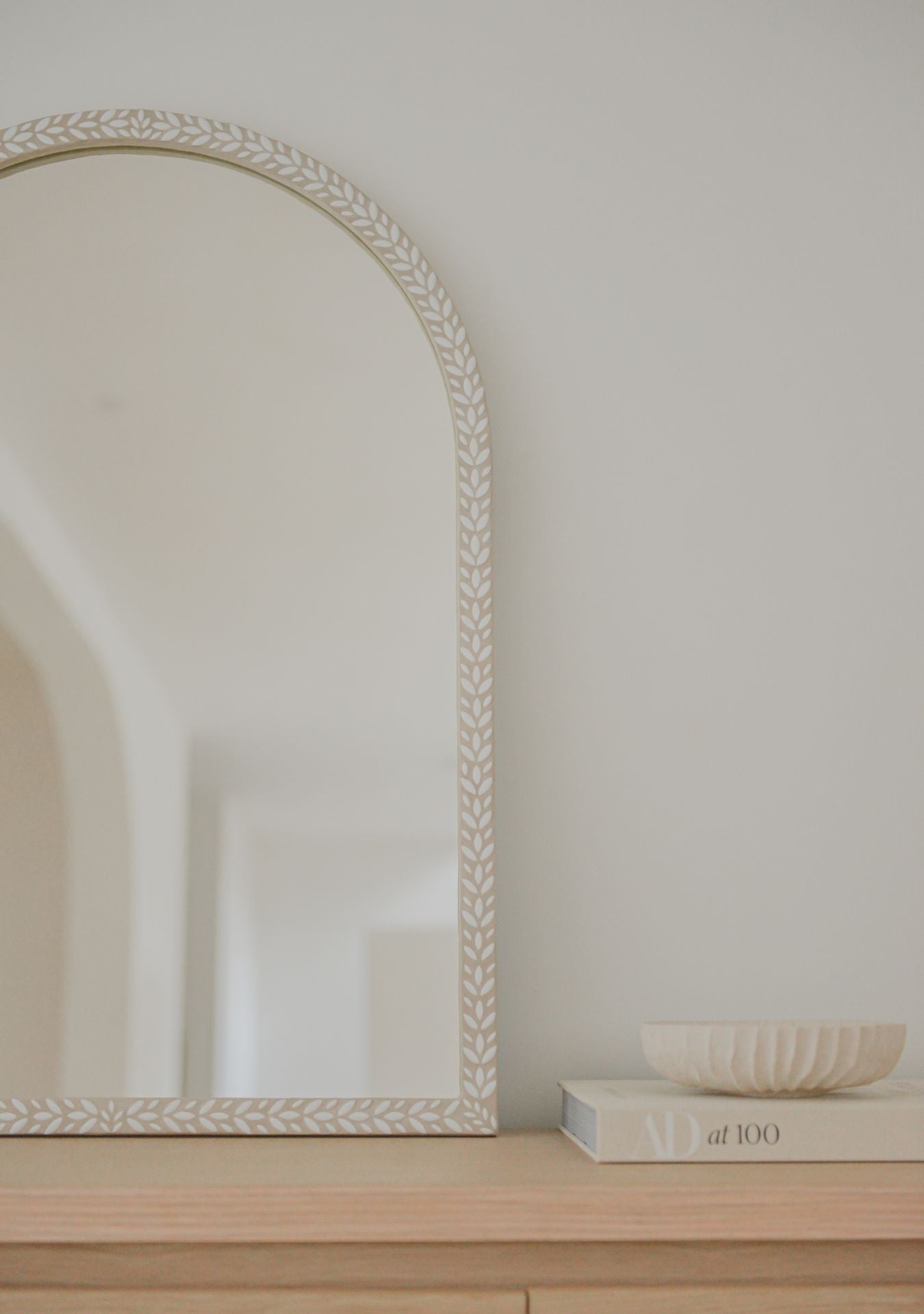 Close-up of a handmade white arched mosaic mirror by Pure Reflection, featuring a delicate leaf-patterned frame styled on a light wooden cabinet in a calm, minimal interior.